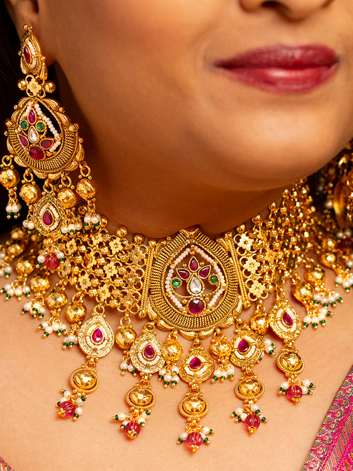A woman in a pink and gold outfit smiles softly, wearing the Zoharika Choker Necklace Set—an elegant gold necklace set in jali work, with matching earrings adorned with red and white colored stones, ghungroo stones and more. Perfect for wedding outfits and the festive season. Blurred green foliage forms the background.