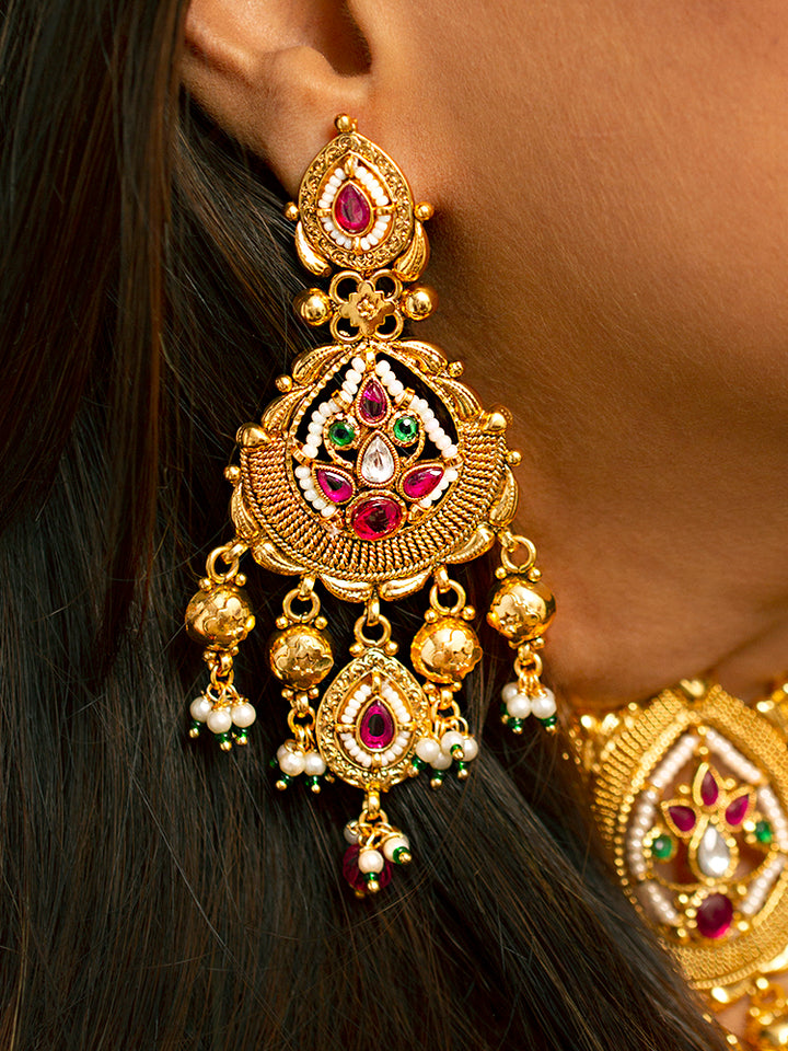 A woman in a pink and gold outfit smiles softly, wearing the Zoharika Choker Necklace Set—an elegant gold necklace set in jali work, with matching earrings adorned with red and white colored stones, ghungroo stones and more. Perfect for wedding outfits and the festive season. Blurred green foliage forms the background.