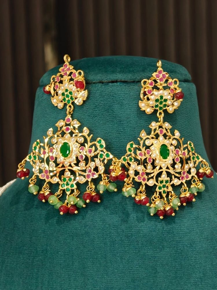 A pair of ornate gold-toned jadau Indian chandbali earrings decorated with green and red stones and hanging bead drops, displayed on a dark green velvet jewelry stand.