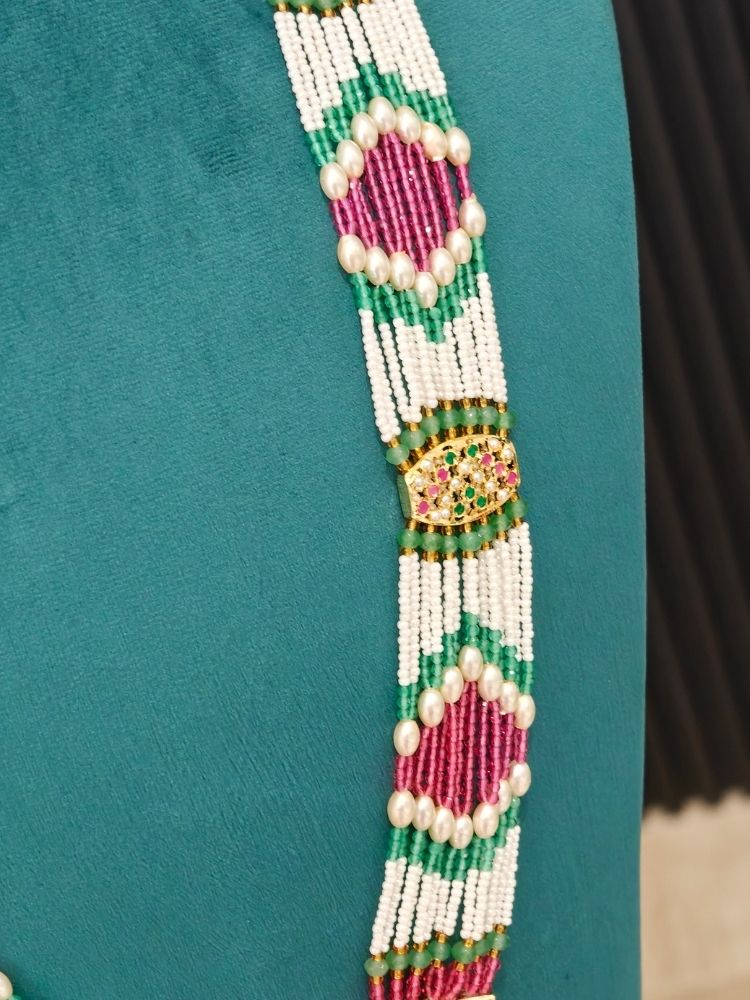 A close-up of a jadau necklace featuring a vertical decorative beaded panel with white beads, pink and green geometric patterns, and a central gold-toned embellished motif.