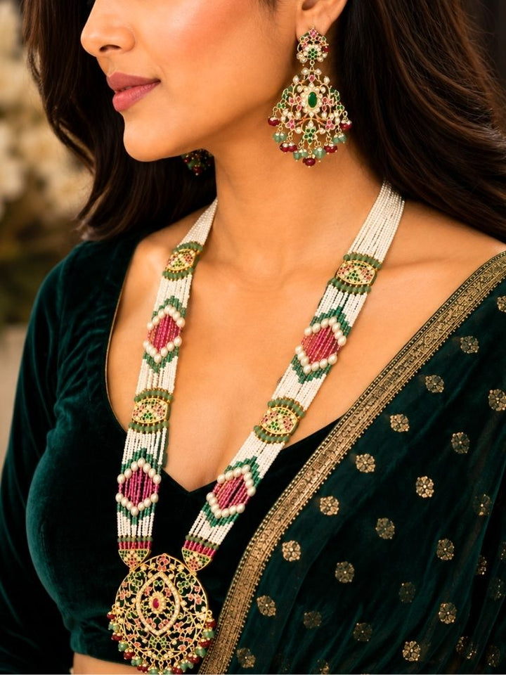 A close-up lifestyle shot of a woman wearing an ornate traditional Indian jadau jewelry set, featuring a long beaded necklace with a large pendant and matching drop earrings. She is wearing a dark green velvet blouse with gold embroidery and a matching embroidered saree.