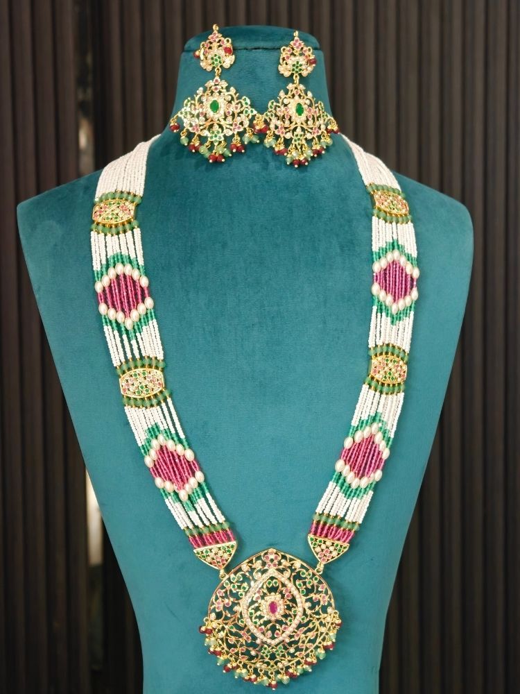 A traditional Indian jadau jewelry set consisting of a statement necklace and matching earrings, displayed on a teal velvet mannequin bust. The jewelry features a white beaded base with intricate gold-plated filigree, green and pink stone embellishments, and red bead accents.