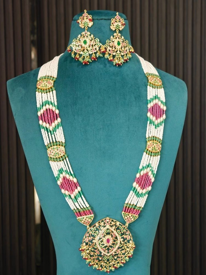 A traditional Indian jadau jewelry set consisting of a statement necklace and matching earrings, displayed on a teal velvet mannequin bust. The jewelry features a white beaded base with intricate gold-plated filigree, green and pink stone embellishments, and red bead accents.