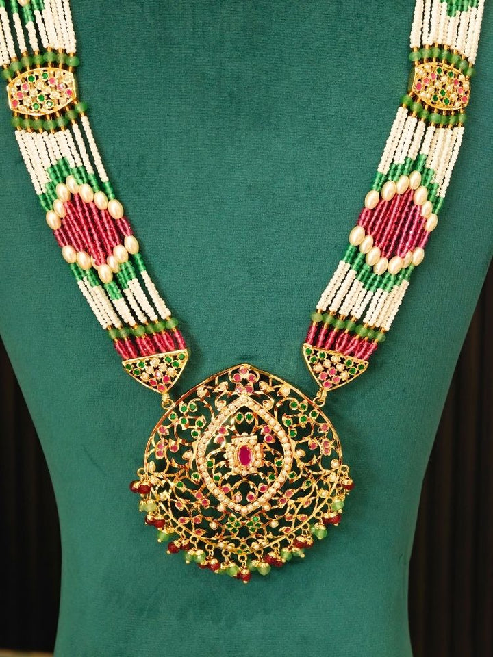 An ornate traditional Indian statement jadau necklace displayed on a dark green velvet mannequin bust. The necklace features a large, intricate gold filigree pendant embellished with red and green stones, connected to a multi-strand beaded chain of white, green, and red beads.