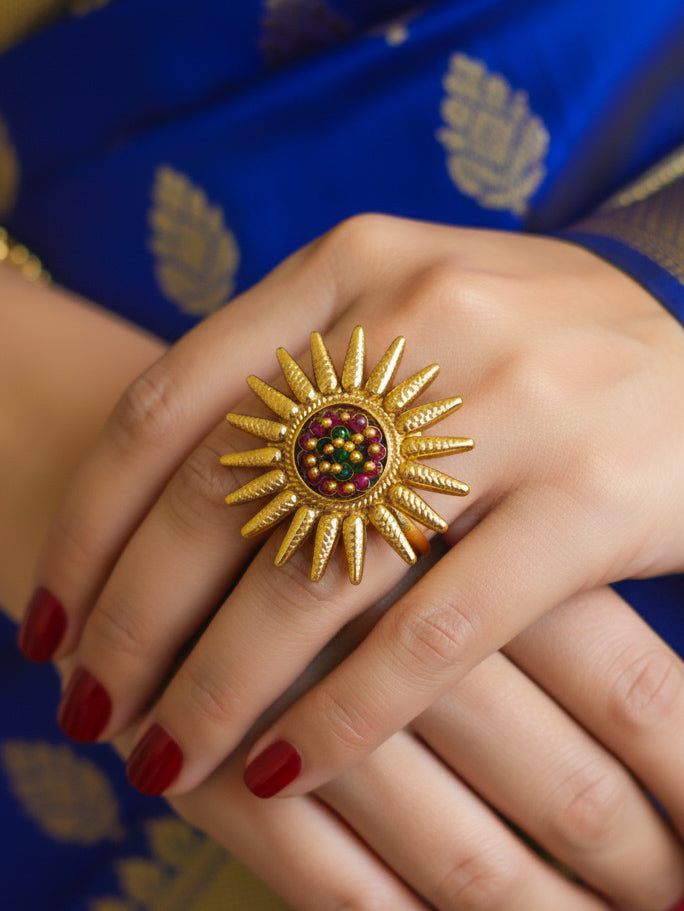 Gold sun-shaped cocktail ring with red and yellow colored stones in the middle on a hand against a blue fabric background. 